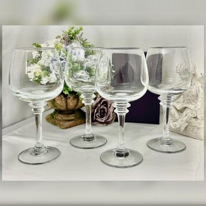 May include: Four clear wine glasses with rounded bowls and long stems. The glasses are arranged on a white surface, with a floral arrangement and decorative elements in the background. The glasses are made of transparent glass.