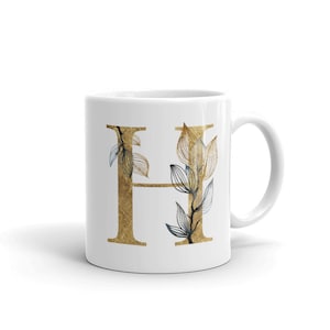 Puede incluir: Taza de cerámica blanca con una letra H dorada y un diseño floral azul y dorado.