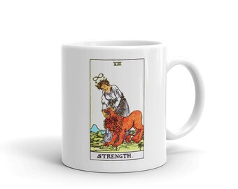 May include: White ceramic mug with a colorful illustration of the Strength tarot card. The card depicts a woman with a crown of flowers, gently holding the mouth of a roaring lion. The card number, VIII, and the word STRENGTH are printed below the image.