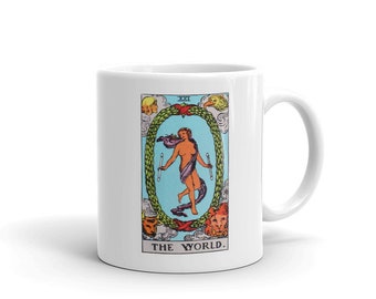 May include: White ceramic mug with a colorful illustration of the World tarot card. The card features a woman standing in a wreath with four animals surrounding her. The text "THE WORLD" is printed below the card.
