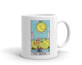May include: White ceramic mug featuring a tarot card design. The card depicts a yellow sun with a face, two dogs, and the text "THE MOON." The design is framed by a blue border with the Roman numeral XVIII at the top.