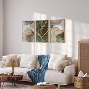 Puede incluir: Tres impresiones de arte abstracto enmarcadas con dise&ntilde;os de hojas doradas y verdes. Las impresiones est&aacute;n colgadas en una pared blanca sobre un sof&aacute; blanco con una manta de tiro azul.