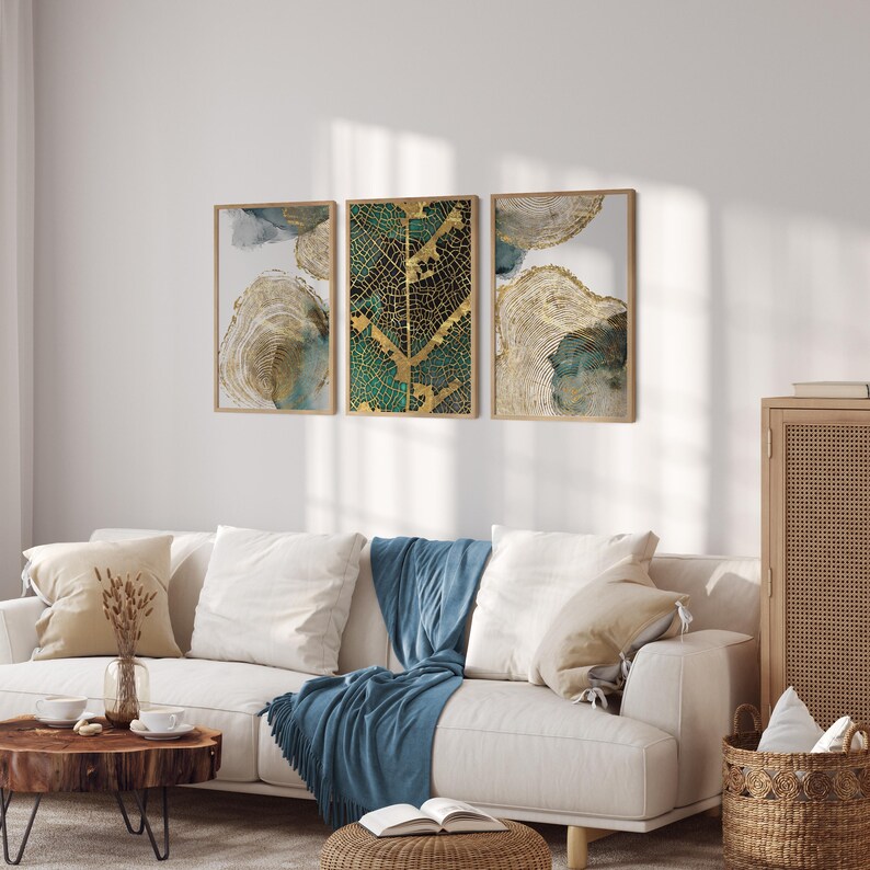 Puede incluir: Tres impresiones de arte abstracto enmarcadas con dise&ntilde;os de hojas doradas y verdes. Las impresiones est&aacute;n colgadas en una pared blanca sobre un sof&aacute; blanco con una manta de tiro azul.