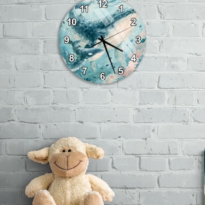 Op de afbeelding: Een ronde wandklok met een aquarelillustratie van een grote walvis en een kleinere walvis die in blauw oceaanwater met koraalriffen zwemmen. De wijzerplaat heeft zwarte cijfers van 1 tot 12 en zwarte uur-, minuut- en secondewijzers.