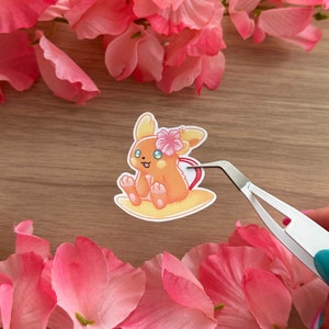 May include: A sticker featuring a cartoon illustration of a small, orange, fox-like Pokemon with a pink flower on its head. The Pokemon is sitting on a yellow surfboard.