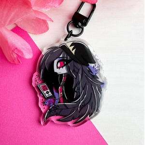 May include: A clear acrylic keychain featuring a cartoon character with long black hair, wearing a black hat and a red shirt. The character is holding a small music player with a play button. The background is a purple and blue gradient with white stars.