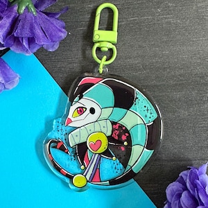 May include: A colorful, cartoon jester character with a green, black, and blue patterned costume. The jester is holding a scepter with a heart on top. The character is on a clear acrylic keychain with a green metal clasp.
