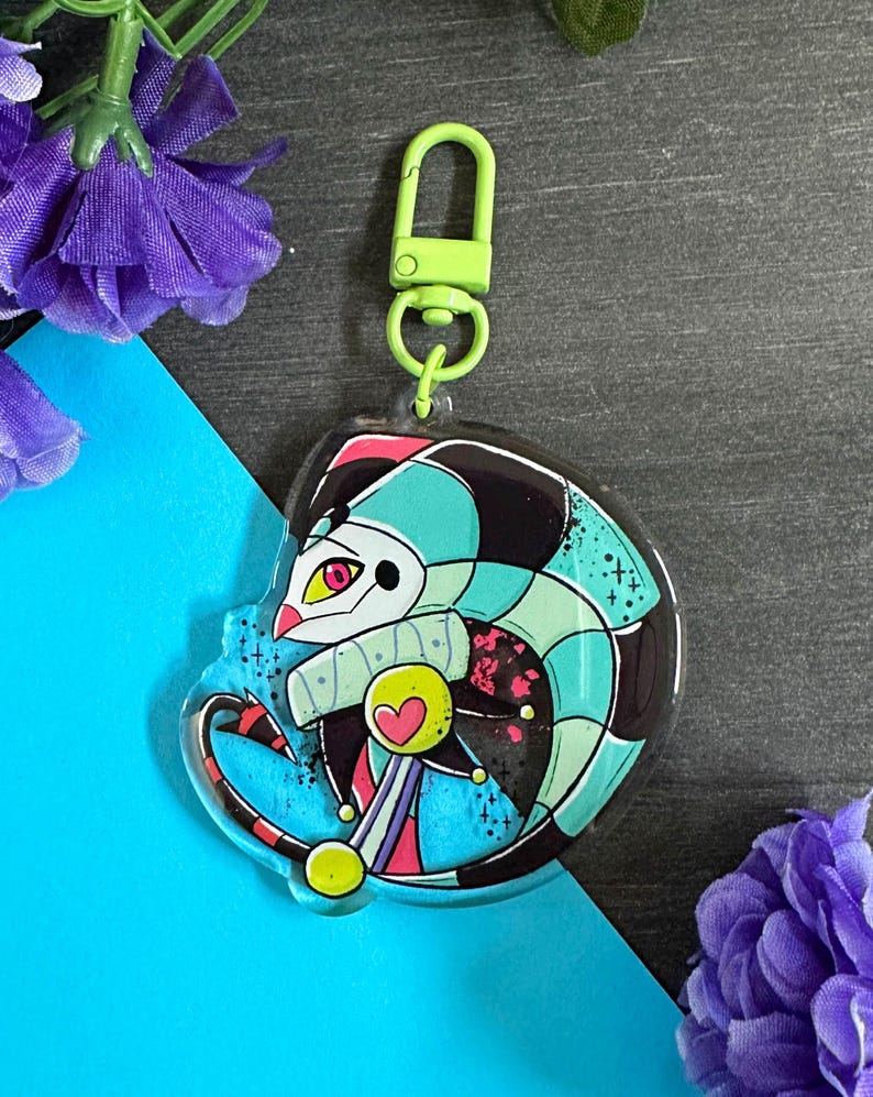 May include: A colorful, cartoon jester character with a green, black, and blue patterned costume. The jester is holding a scepter with a heart on top. The character is on a clear acrylic keychain with a green metal clasp.