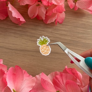May include: A small, yellow pineapple sticker with green leaves is being held by tweezers on a wooden surface. The sticker is surrounded by pink flower petals.