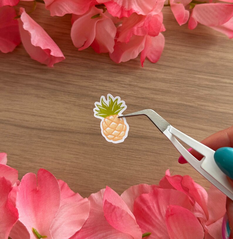 May include: A small, yellow pineapple sticker with green leaves is being held by tweezers on a wooden surface. The sticker is surrounded by pink flower petals.