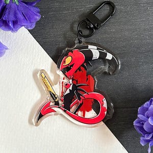 May include: A red and black cartoon character with a sword, holding a red and white striped tail, with blood splatters on a white background. The character is attached to a black keychain.