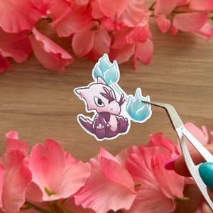 May include: A sticker featuring a cartoon purple and white creature with blue flames. The creature is holding the flames in its hands. The sticker is on a wooden surface.