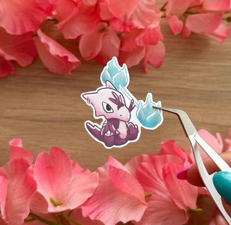 May include: A sticker featuring a cartoon purple and white creature with blue flames. The creature is holding the flames in its hands. The sticker is on a wooden surface.