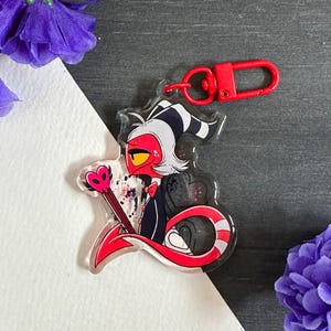 May include: A red and black cartoon character with a white and black striped tail, holding a pink heart-shaped wand. The character is on a clear acrylic keychain with a red metal ring.