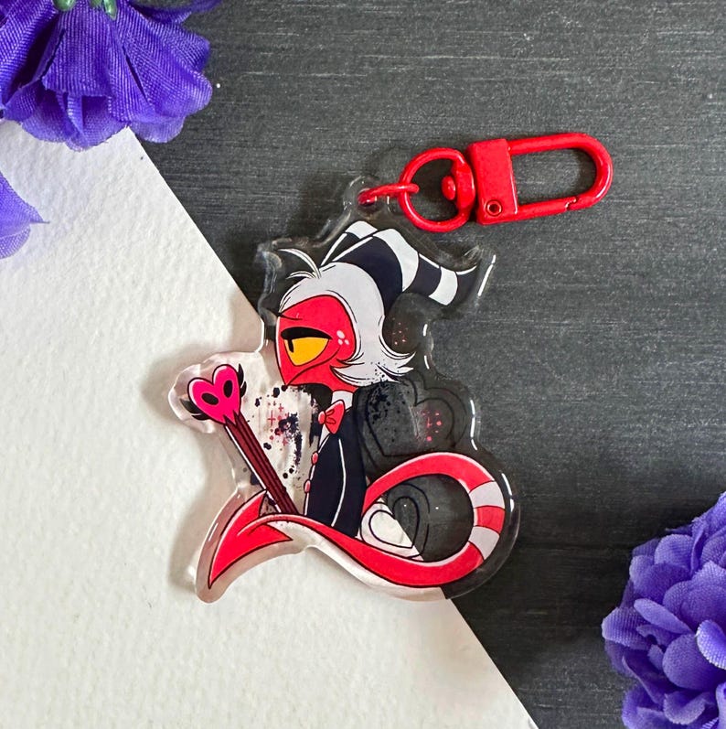 May include: A red and black cartoon character with a white and black striped tail, holding a pink heart-shaped wand. The character is on a clear acrylic keychain with a red metal ring.