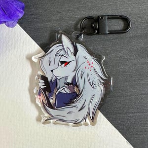 May include: A white and gray anime character with red eyes and black hair holding a black phone. The character is on a clear acrylic keychain with a black metal ring.
