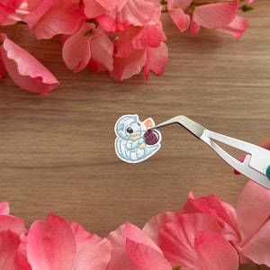 May include: A white and grey cartoon sticker of a small, curled-up creature holding a red berry. The sticker is being held by tweezers.