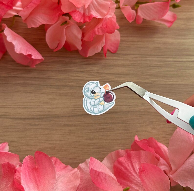 May include: A white and grey cartoon sticker of a small, curled-up creature holding a red berry. The sticker is being held by tweezers.