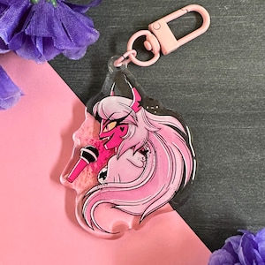 May include: Pink and black anime-style character keychain with a microphone. The character has pink hair, red eyes, and horns. The keychain is made of clear acrylic and has a pink glitter background.