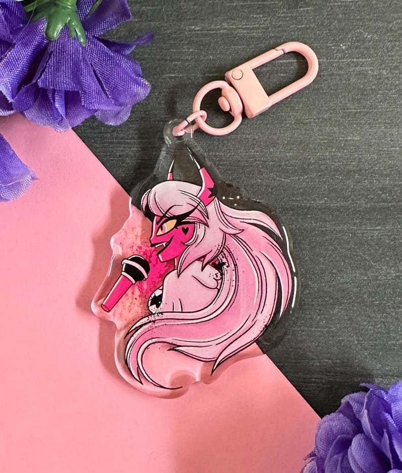 May include: Pink and black anime-style character keychain with a microphone. The character has pink hair, red eyes, and horns. The keychain is made of clear acrylic and has a pink glitter background.