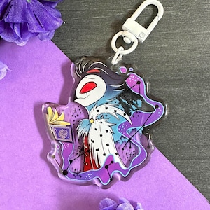 May include: A purple, blue, and white cartoon character with a crown and a book in their hand. The character is surrounded by a purple and blue nebula with stars. The character is on a clear acrylic keychain with a white metal clasp.