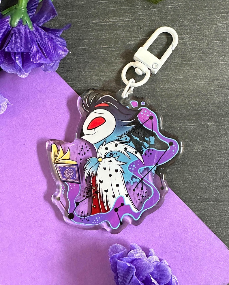May include: A purple, blue, and white cartoon character with a crown and a book in their hand. The character is surrounded by a purple and blue nebula with stars. The character is on a clear acrylic keychain with a white metal clasp.