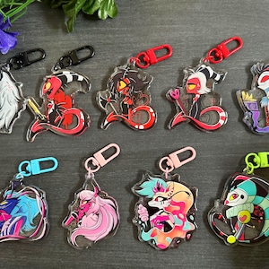 May include: Eight colorful keychains featuring cartoon characters from the animated series Helluva Boss. The keychains are made of clear acrylic and feature a variety of characters, including a red demon with horns, a blue demon with wings, and a pink demon with a tail.
