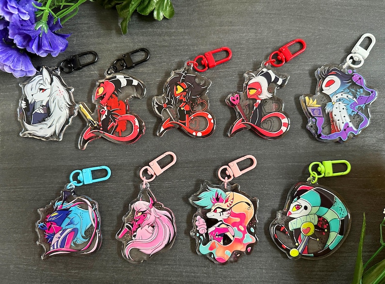 May include: Eight colorful keychains featuring cartoon characters from the animated series Helluva Boss. The keychains are made of clear acrylic and feature a variety of characters, including a red demon with horns, a blue demon with wings, and a pink demon with a tail.