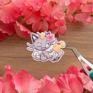 May include: A sticker featuring a cartoon purple cat with a winking eye holding a yellow pineapple drink with a pink flower on top. The sticker is on a wooden surface.