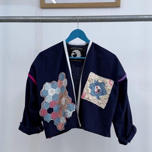 May include: A navy blue cropped jacket with a white trim and a patchwork design on the front. The patchwork includes a hexagonal pattern in shades of blue, pink, and white, and a square patch in shades of blue and white. The jacket has a label that reads "MAYER PIECE" with a stylized bird logo.