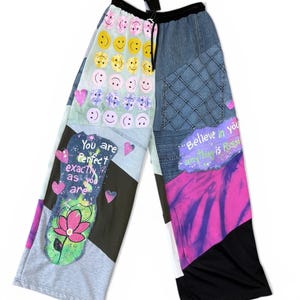 May include: Patchwork trousers with a black drawstring waist. The design includes denim, tie-dye, and grey panels, adorned with smiley faces, hearts, and text that reads "You are perfect exactly as you are" and "Believe in you anything is possible."