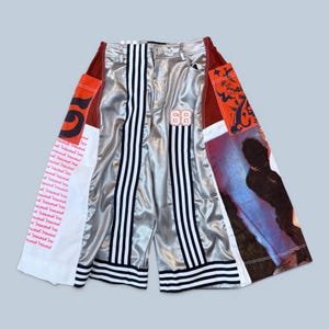 Pode incluir: Calções desportivos prateados com riscas brancas e pretas e aplicações de tecido laranja, branco e vermelho. Os calções têm o número "68" e um design de riscas brancas e pretas.