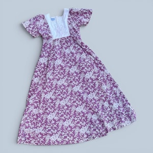 Puede incluir: Un vestido largo de estilo vintage con estampado floral. El vestido presenta un fondo morado oscuro con detalles florales blancos. Tiene mangas cortas abullonadas y un escote cuadrado con detalles bordados en blanco.
