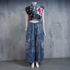 May include: A cropped denim vest featuring various patches, including a large red number 41, worn over a white long-sleeve shirt. Paired with wide-leg blue denim trousers that have a button-down front, multiple pockets, and frayed edges.