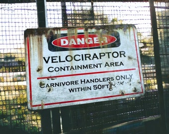 May include: A red and white warning sign with the text "DANGER VELOCIRAPTOR CONTAINMENT AREA CARNIVORE HANDLERS ONLY WITHIN 50FT" on a rusty metal background.