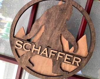 May include: A wooden round ornament with a silhouette of Bigfoot walking in front of mountains. The ornament is brown and has the name "Schaffer" engraved on it.
