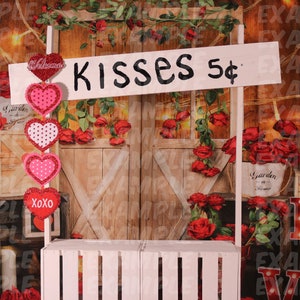 Könnte beinhalten: Ein Fotohintergrund zum Thema Valentinstag mit einem Holzschild, auf dem "Kisses 5¢" steht. Der Hintergrund zeigt eine rustikale Holztür mit roten Rosen und Ranken, eine weiße Holzkiste und ein Schild mit der Aufschrift "XoXo".
