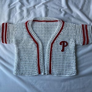 May include: White crochet baseball jersey with red trim and a red "P" patch. The jersey has short sleeves with red stripes and a V-neck design. The open weave creates a breathable, lightweight feel.