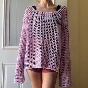 May include: A long-sleeved, loose-fitting sweater crafted from light purple yarn in an open, grid-like crochet pattern. The garment features a wide, rounded neckline and a boxy silhouette, showcasing a textured, semi-sheer construction throughout.