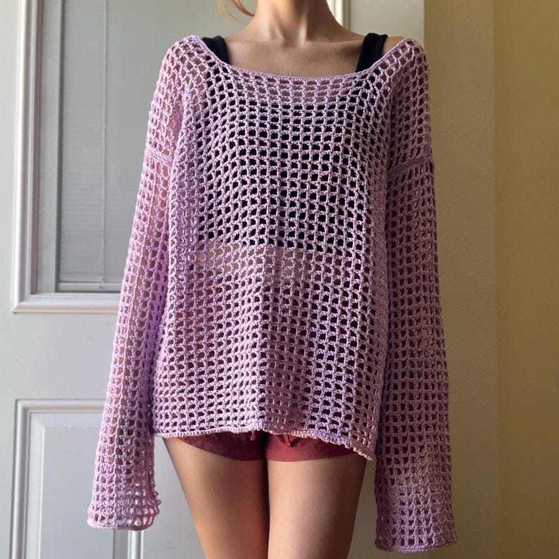 May include: A long-sleeved, loose-fitting sweater crafted from light purple yarn in an open, grid-like crochet pattern. The garment features a wide, rounded neckline and a boxy silhouette, showcasing a textured, semi-sheer construction throughout.