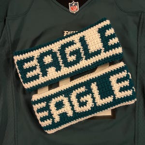 May include: Two handmade crochet headbands with the word "EAGLE" in white letters on a teal background. The headbands are displayed on a green football jersey.