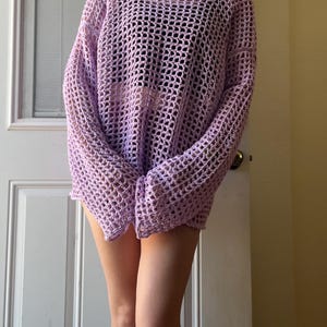 May include: A light purple long-sleeve sweater featuring an open-knit crochet construction with a wide, square neckline and a loose, relaxed fit. The garment is crafted from yarn in a soft lavender hue, showcasing a grid-like pattern throughout.