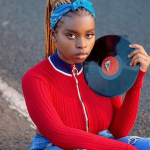 May include: A woman wearing a red ribbed jumper with a blue and white trim, blue jeans, and a blue bandana. She is holding a black vinyl record.