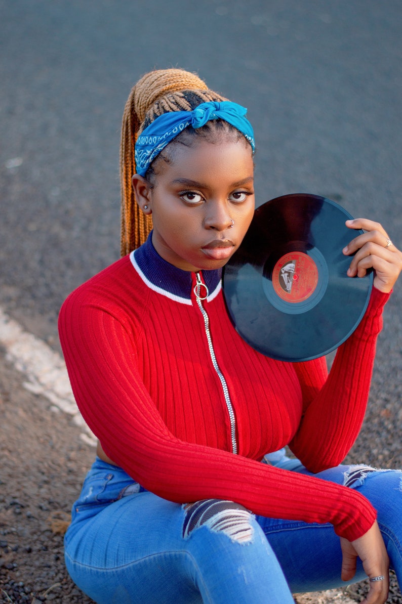 May include: A woman wearing a red ribbed jumper with a blue and white trim, blue jeans, and a blue bandana. She is holding a black vinyl record.