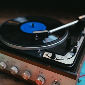 May include: A vintage record player with a black vinyl record on the turntable. The record player has a wooden case with silver and black controls. The record player has a red light that is on.