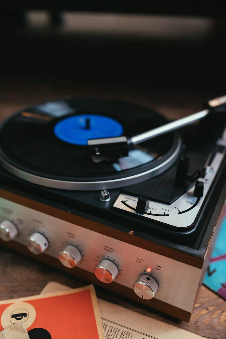 May include: A vintage record player with a black vinyl record on the turntable. The record player has a wooden case with silver and black controls. The record player has a red light that is on.