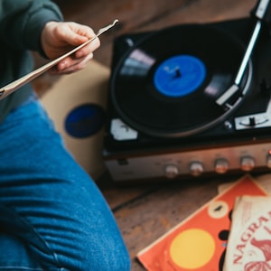 May include: A person is holding a vinyl record in their hands, sitting next to a vintage record player with a black turntable and a blue record on it. There are two additional vinyl records on the wooden floor, one with a yellow circle and the other with a palm tree design and the text "Distillerie Nagra".