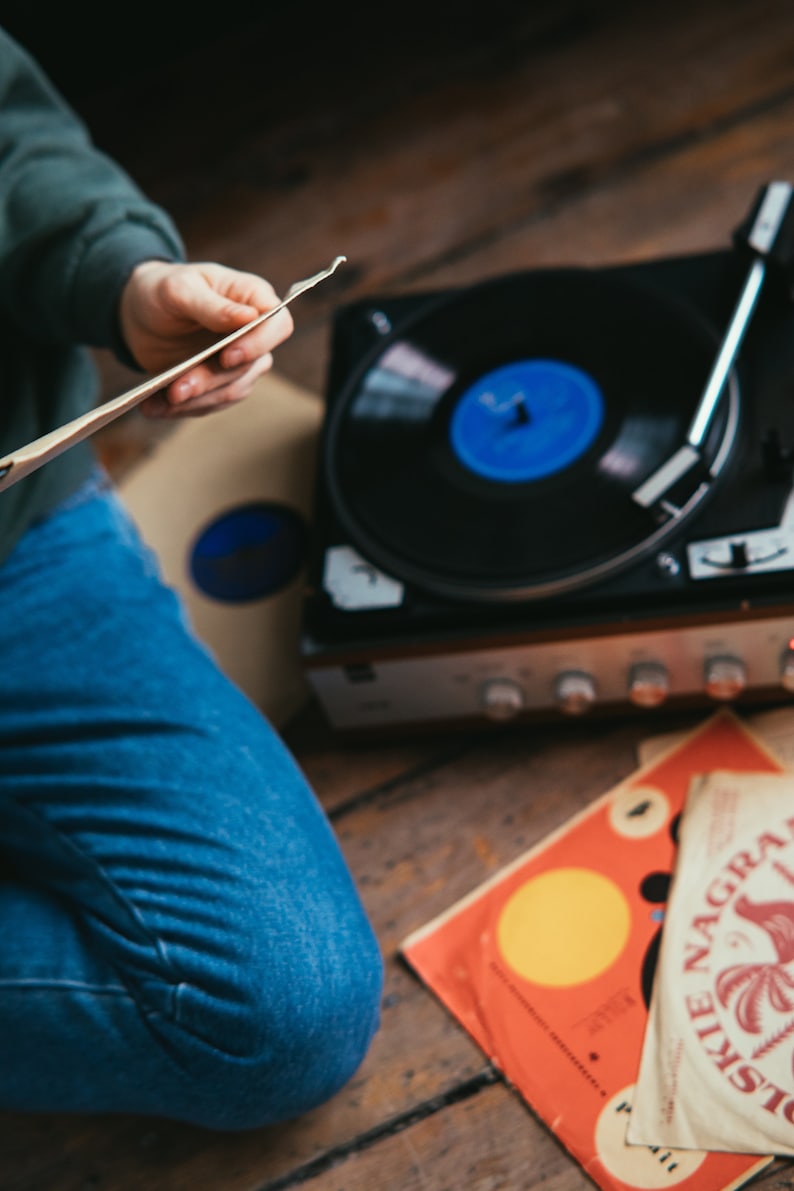 May include: A person is holding a vinyl record in their hands, sitting next to a vintage record player with a black turntable and a blue record on it. There are two additional vinyl records on the wooden floor, one with a yellow circle and the other with a palm tree design and the text "Distillerie Nagra".