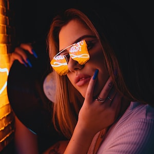 May include: A woman wearing reflective sunglasses with a neon sign reflected in the lenses. She is holding a black vinyl record and has blue nail polish.