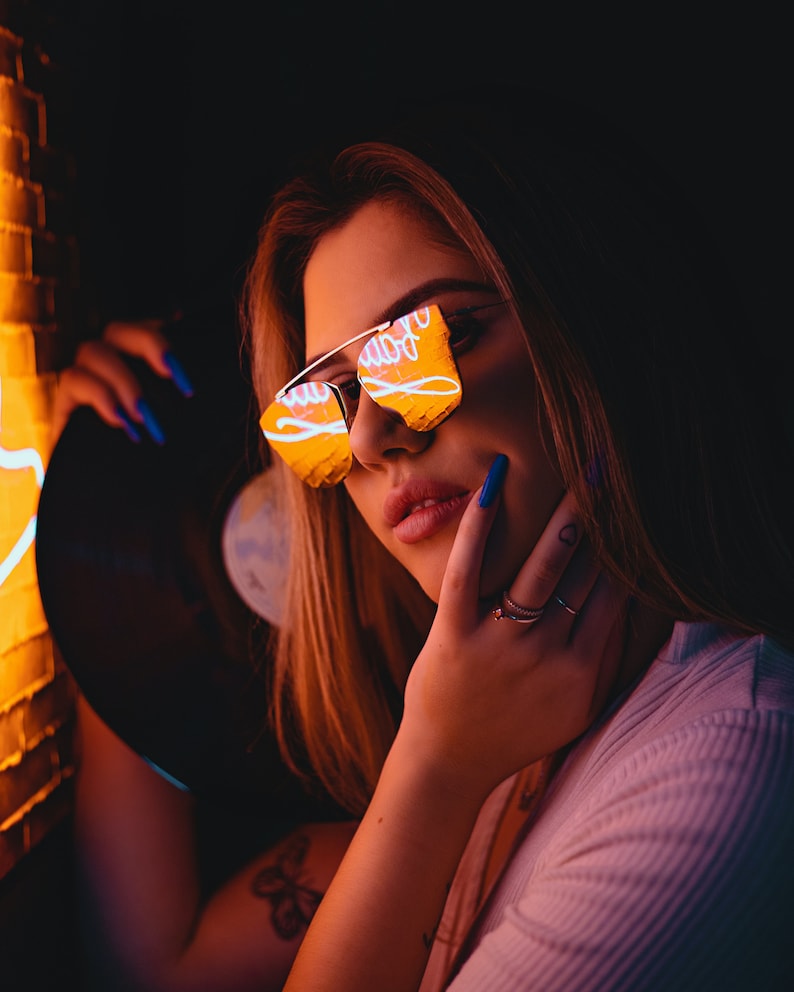 May include: A woman wearing reflective sunglasses with a neon sign reflected in the lenses. She is holding a black vinyl record and has blue nail polish.